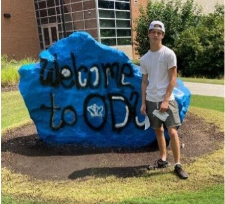 Teenage boy in front of a big blue painted rock in front of a high school