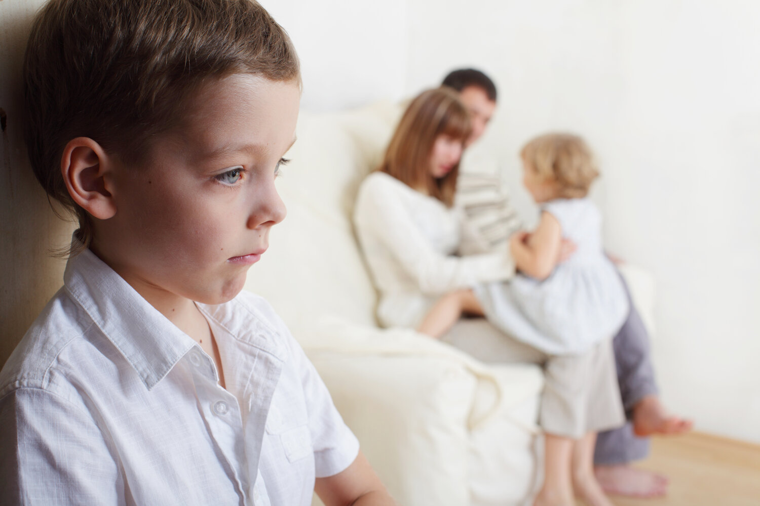 Parents on a white couch with young daughter on their laps. Young brother is separate and frowning signifying being jealous of his younger sister.