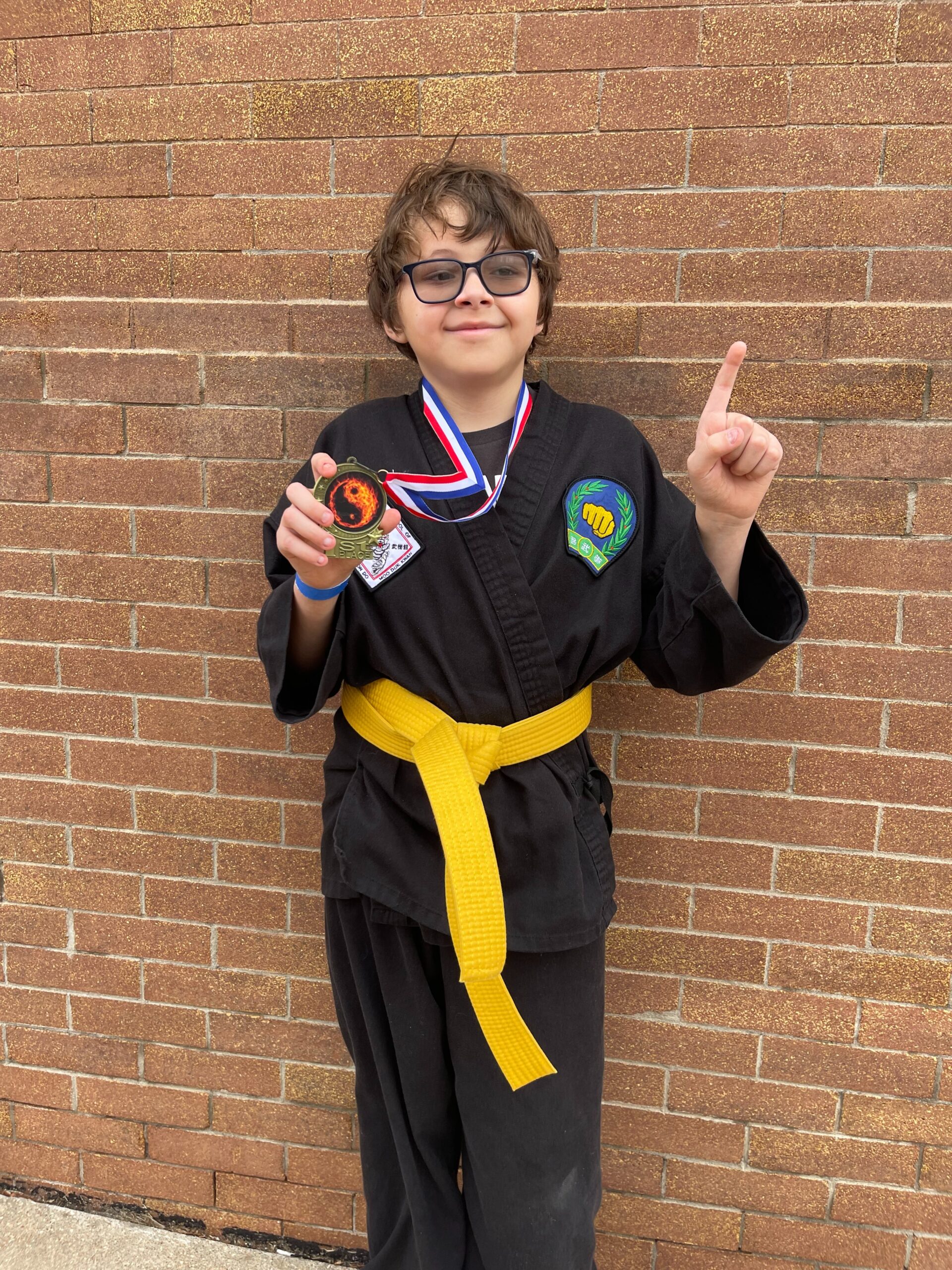 Smiling boy in a black karate uniform with a yellow belt holding a medal and pointing one finger in the air