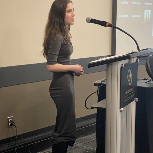 Visually impaired young woman in a grey dress standing a podium and speaking into a microphone
