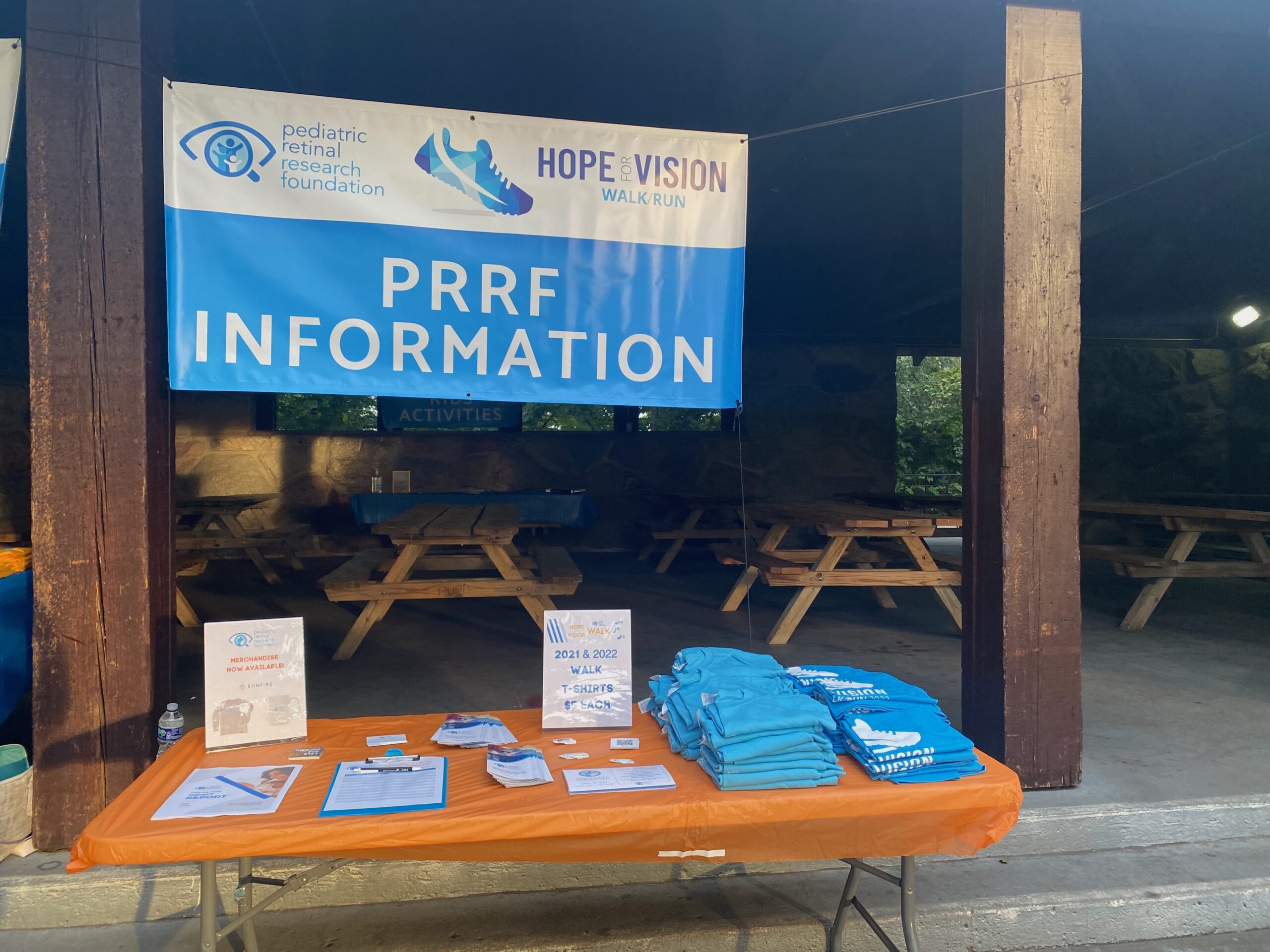 PRRF Hope for Vision Walk & Run Information table with signs, brochures and t-shirts and banner hanging in a park pavilion
