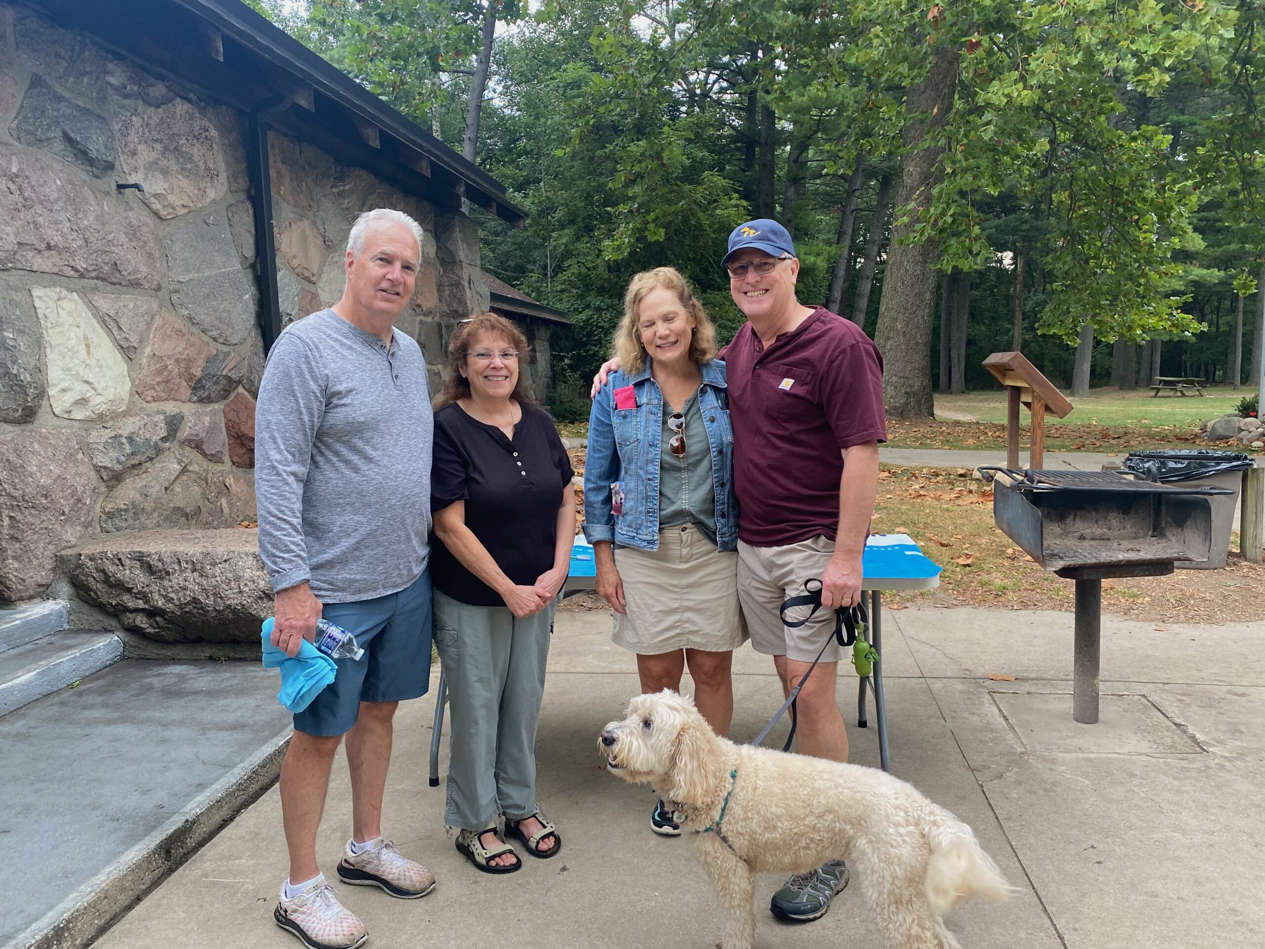 Two smiling older couples and white curly haired dog standing in a park pavilion