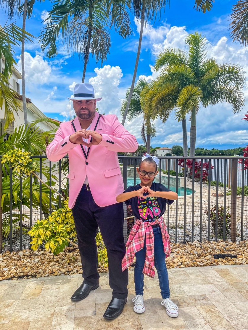Father and young daughter standing in front of a swimming pool and palm trees making a heart with their hands