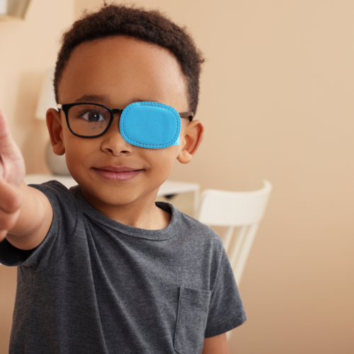 Young boy with glasses and a blue eye patch on one side smiles and gives a thumbs up