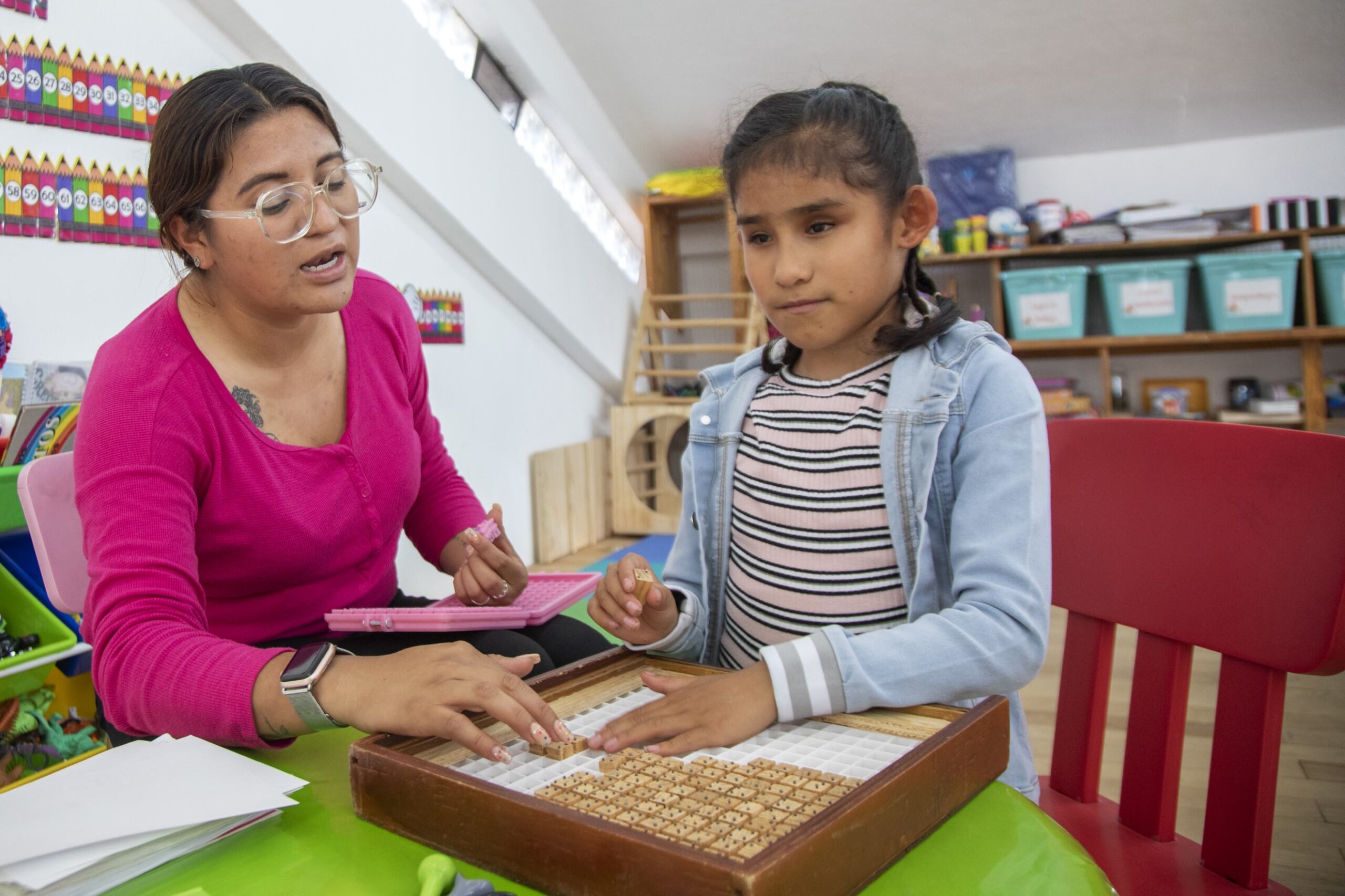 Female teacher with glasses assisting a young female visually impaired student with a wood braille device