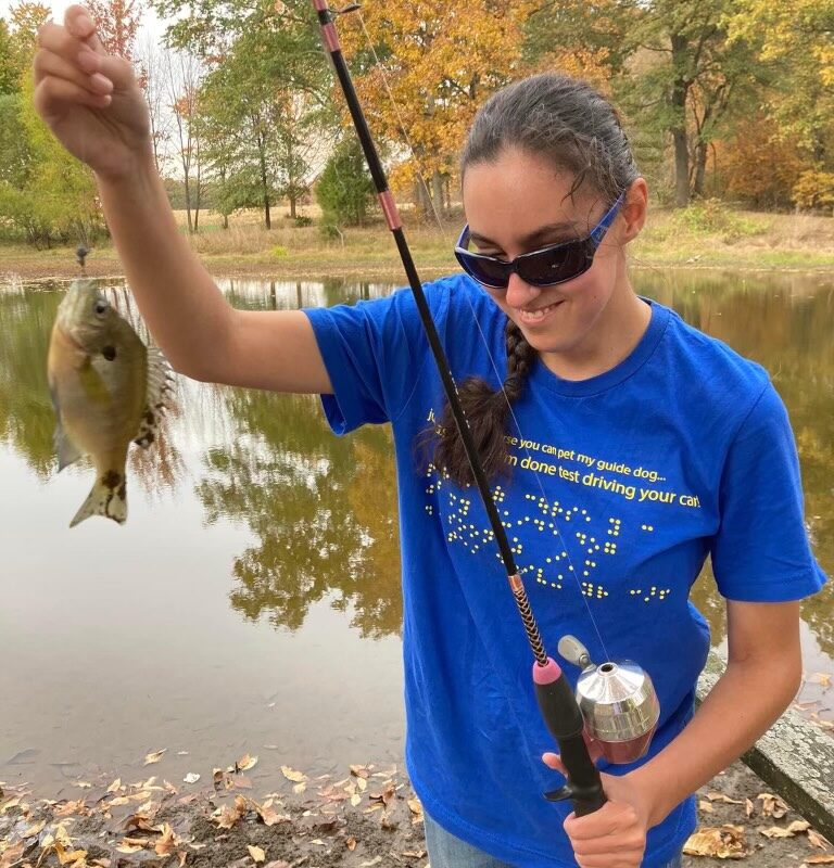 Smiling young woman with sunglasses, a blue t-shirt and jeans holding a fishing rod and a fish at the end of the line