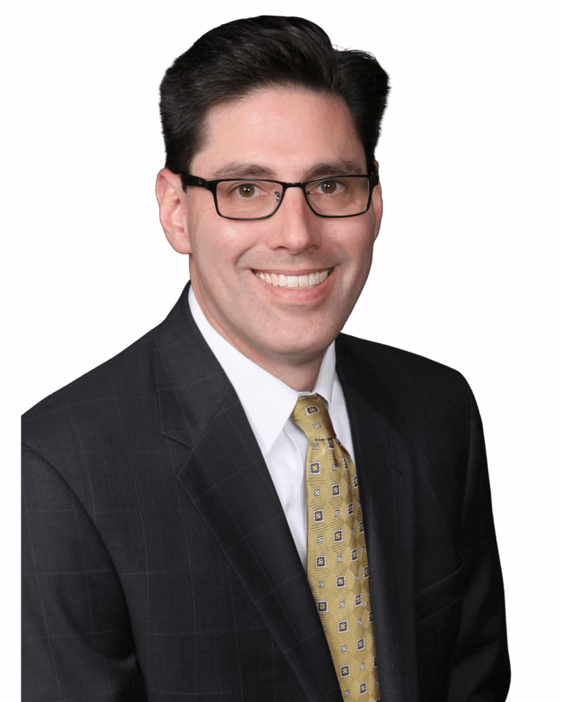 Photo of Adam Zuckerman wearing a black suit and yellow tie, smiling at the camera with glasses on.