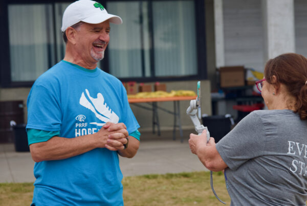 A smiling Dr. Capone wears a bright blue Hope for Vision walk shirt and a white hat. He stands across from an individual in a gray shirt who films him on an iPhone gimbal for a social media interview.