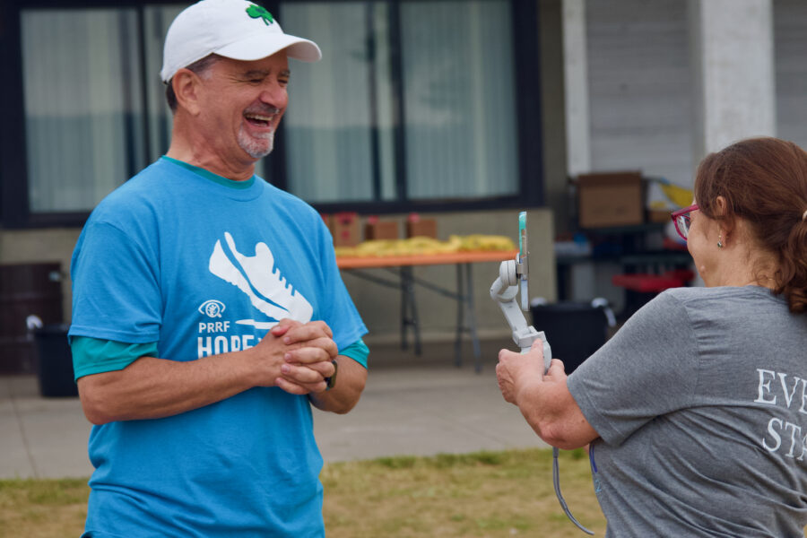 A smiling Dr. Capone wears a bright blue Hope for Vision walk shirt and a white hat. He stands across from an individual in a gray shirt who films him on an iPhone gimbal for a social media interview.