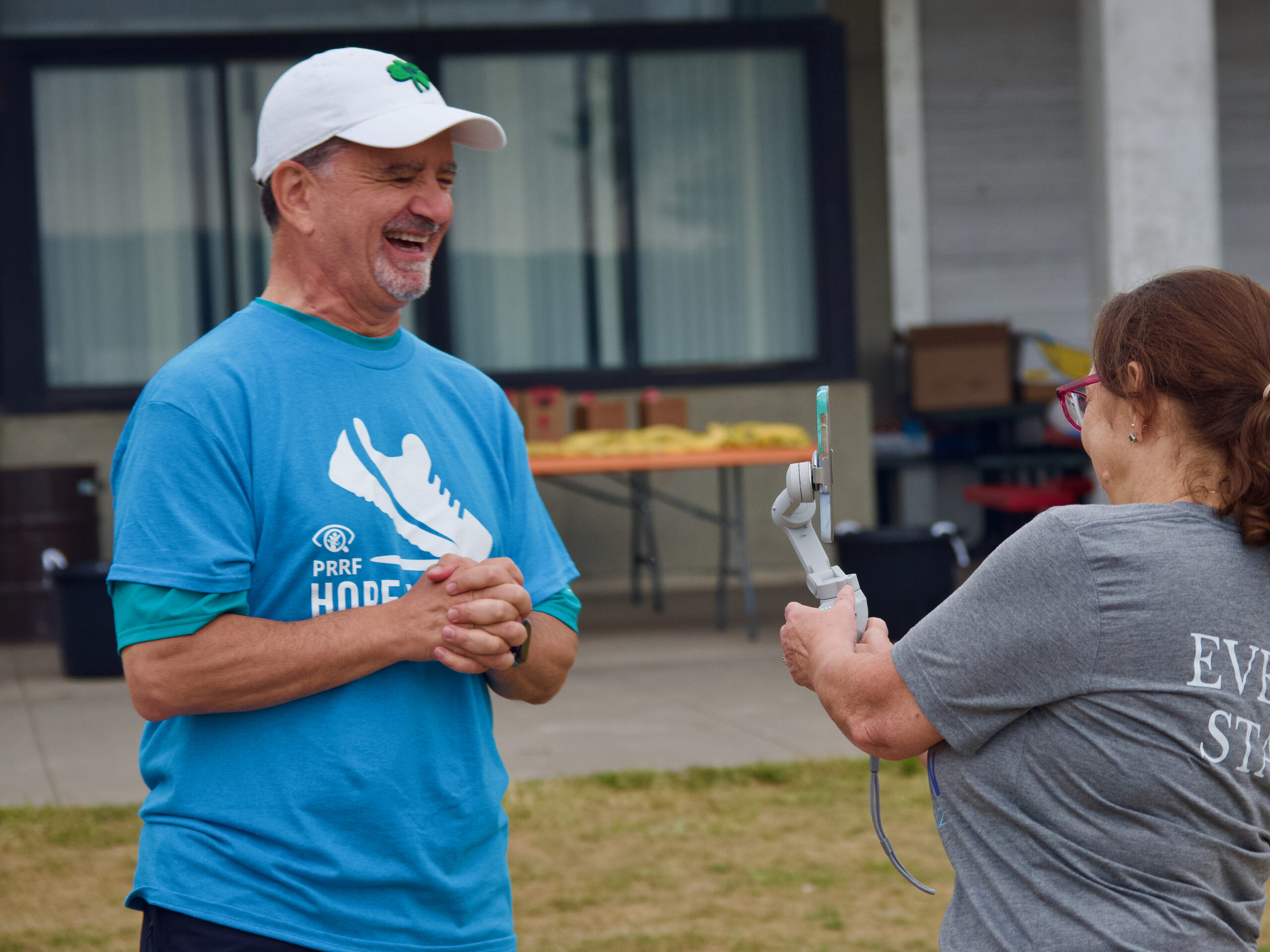 A smiling Dr. Capone wears a bright blue Hope for Vision walk shirt and a white hat. He stands across from an individual in a gray shirt who films him on an iPhone gimbal for a social media interview.