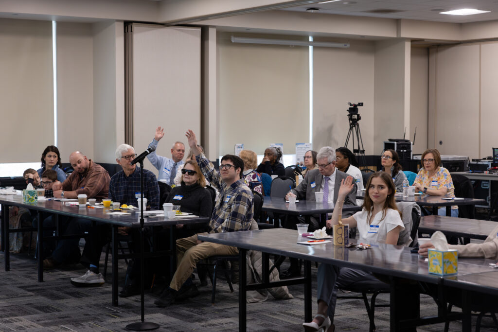 Room in Oakland University with two rows of tables, individuals sitting behind tables in various professional attire have their hands raised.