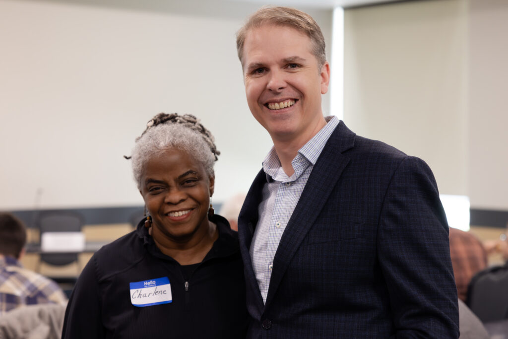 An individual with a dark shirt and name tag reading Charlene stands next to another individual wearing a suit. They are both smiling warmly. The atmosphere is professional.