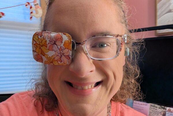 A close-up selfie of Emilee smiling warmly at the camera. She has curly light brown hair pulled back and is wearing clear-framed eyeglasses. Over the right lens of her glasses is a fabric eye patch featuring a retro pattern of orange, yellow, and pink flowers. She is wearing a coral pink crewneck t-shirt, a silver toggle chain necklace, and a small beaded necklace. In the background is a bright room with pink walls, a window with white blinds, and a computer monitor.