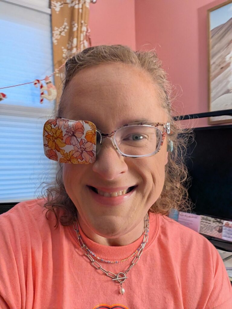 A close-up selfie of Emilee smiling warmly at the camera. She has curly light brown hair pulled back and is wearing clear-framed eyeglasses. Over the right lens of her glasses is a fabric eye patch featuring a retro pattern of orange, yellow, and pink flowers. She is wearing a coral pink crewneck t-shirt, a silver toggle chain necklace, and a small beaded necklace. In the background is a bright room with pink walls, a window with white blinds, and a computer monitor.