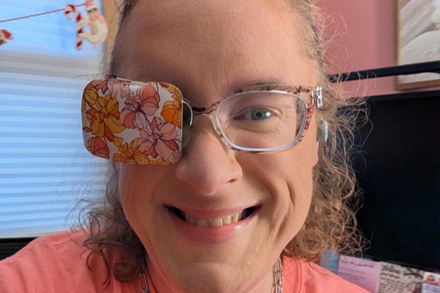 A close-up selfie of Emilee smiling warmly at the camera. She has curly light brown hair pulled back and is wearing clear-framed eyeglasses. Over the right lens of her glasses is a fabric eye patch featuring a retro pattern of orange, yellow, and pink flowers. She is wearing a coral pink crewneck t-shirt, a silver toggle chain necklace, and a small beaded necklace. In the background is a bright room with pink walls, a window with white blinds, and a computer monitor.