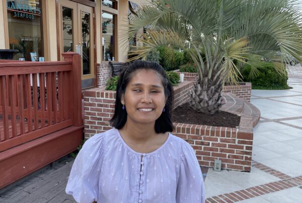 Taengkwa Marie Sturgell, PRRF’s newest volunteer blog writer, smiling outdoors in front of a restaurant with a lit marquee sign and a brick planter with a palm tree. She is wearing a lavender puff-sleeve blouse and blue jeans, with small purple earrings and a blue wristband. The setting appears to be a warm-weather outdoor dining or entertainment district.