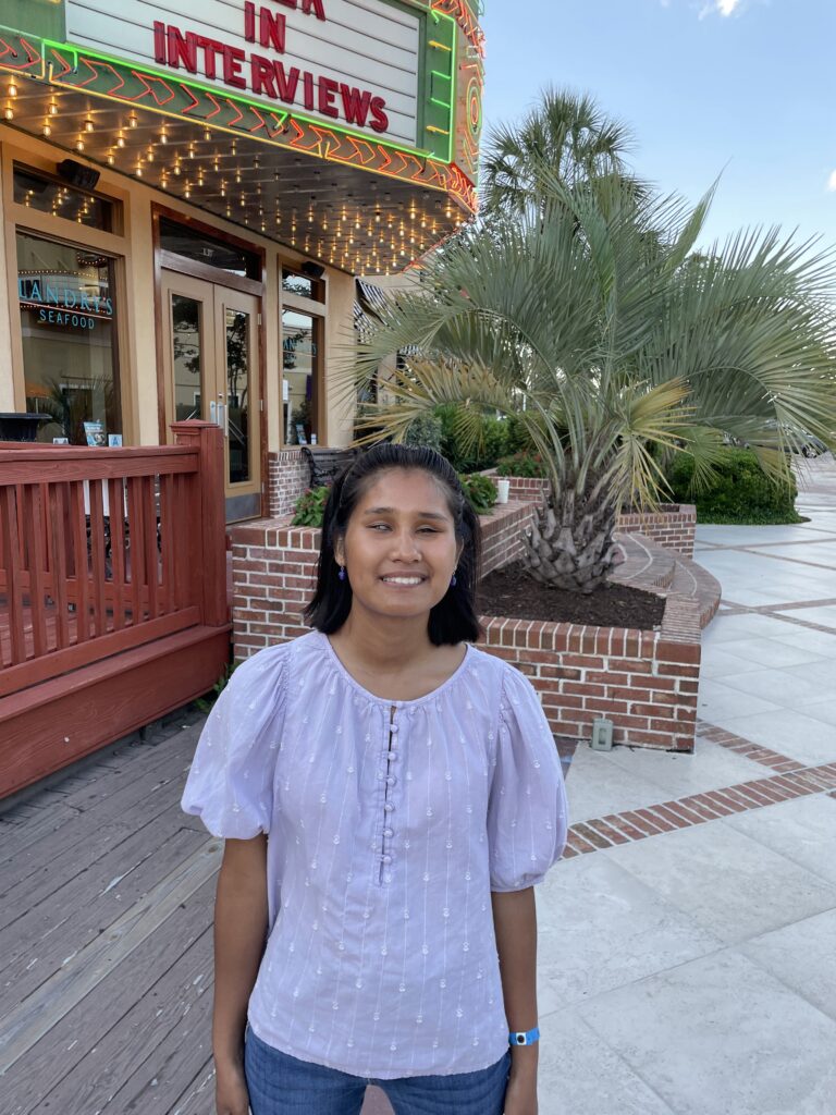 Taengkwa Marie Sturgell, PRRF’s newest volunteer blog writer, smiling outdoors in front of a restaurant with a lit marquee sign and a brick planter with a palm tree. She is wearing a lavender puff-sleeve blouse and blue jeans, with small purple earrings and a blue wristband. The setting appears to be a warm-weather outdoor dining or entertainment district.
